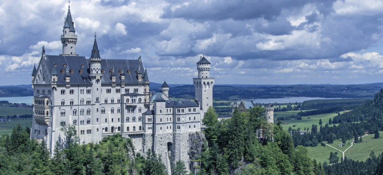 A panoramic view of Neuschwanstein Castle in Bavaria, Germany, surrounded by lush forests and distant lakes under a dramatic cloudy sky.